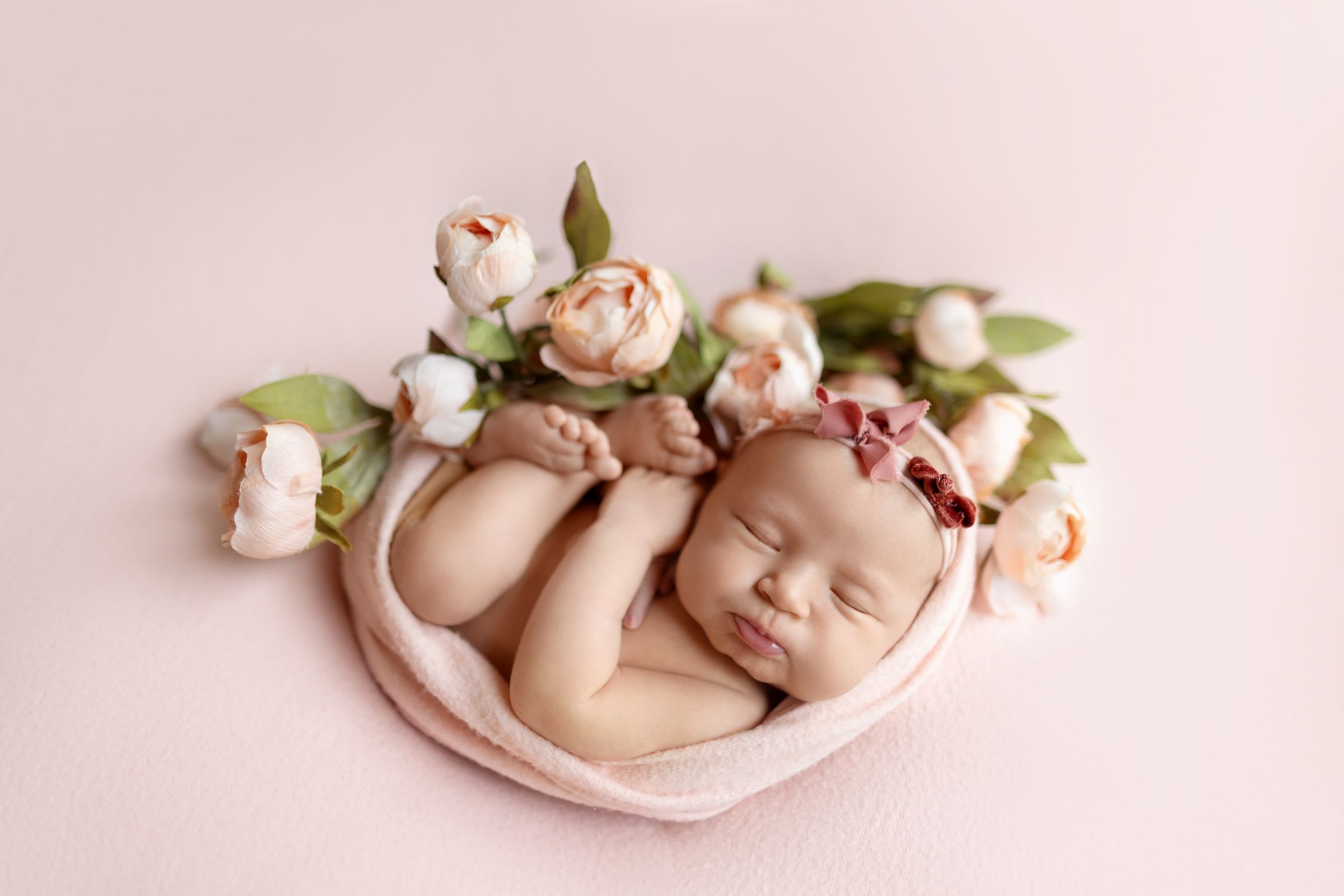 Delicate newborn photography series featuring soft pink tones, floral arrangements, and curled sleeping poses on fabric.
