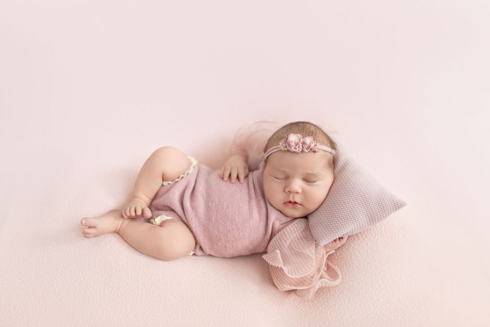 Sleeping newborn baby girl in pink wrap and floral headband resting on a small pillow on a soft pink backdrop.
