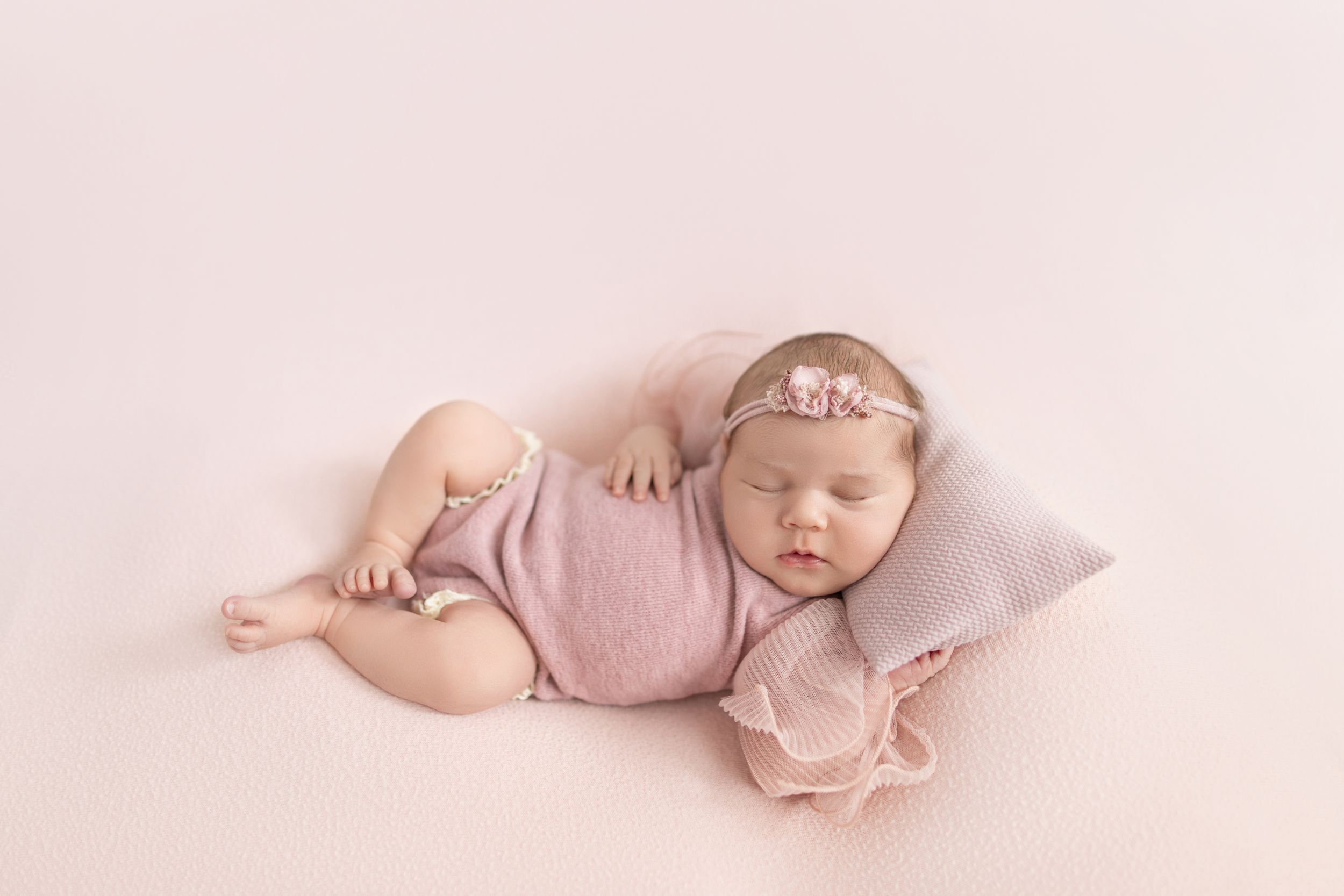 Sleeping newborn baby girl in pink wrap and floral headband resting on a small pillow on a soft pink backdrop.