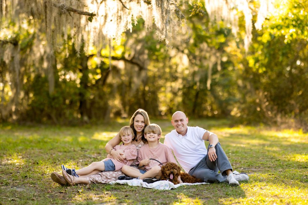 Family of four with a dog sitting on a blanket outdoors under Spanish moss-draped trees.