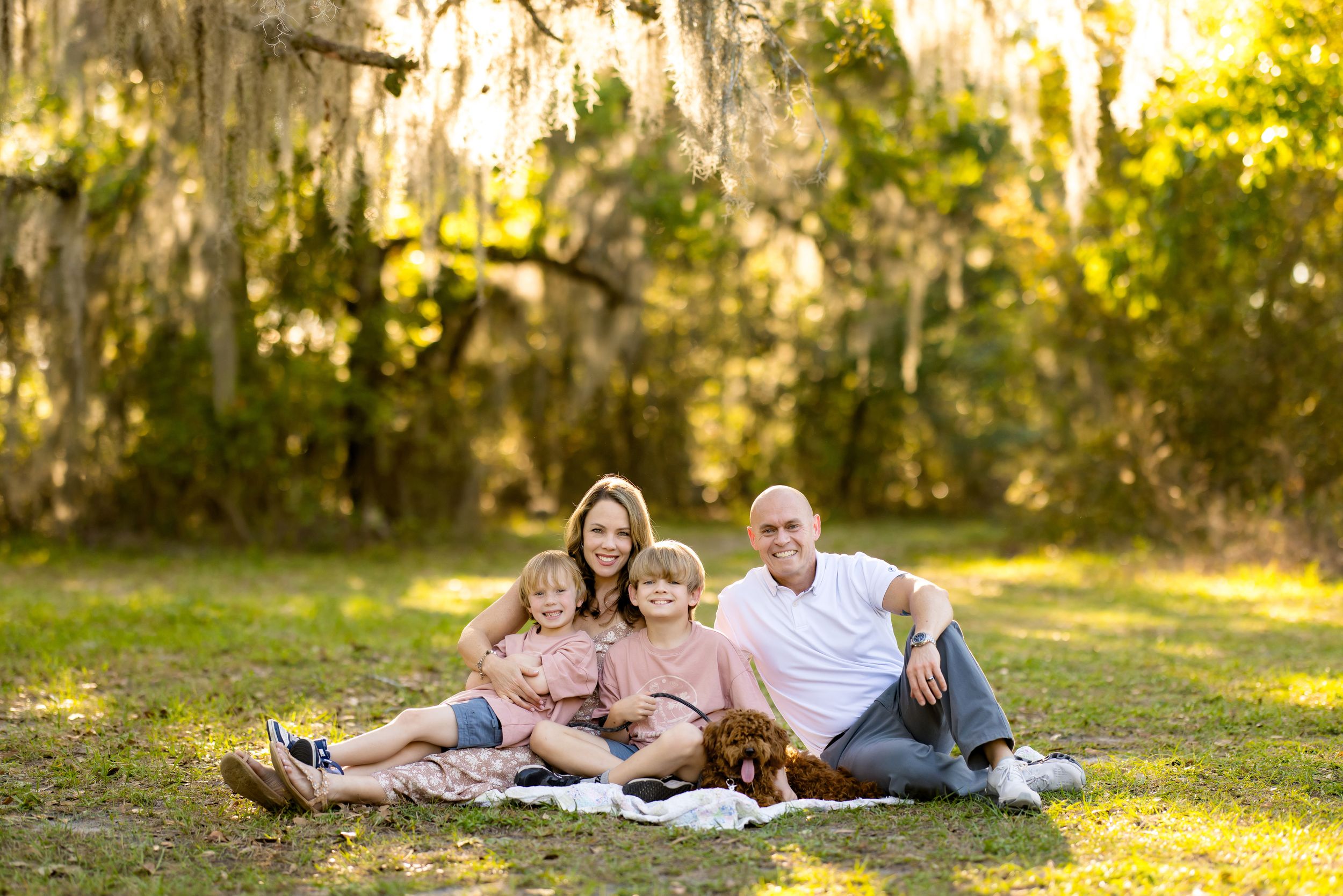 Family of four with a dog sitting on a blanket outdoors under Spanish moss-draped trees.