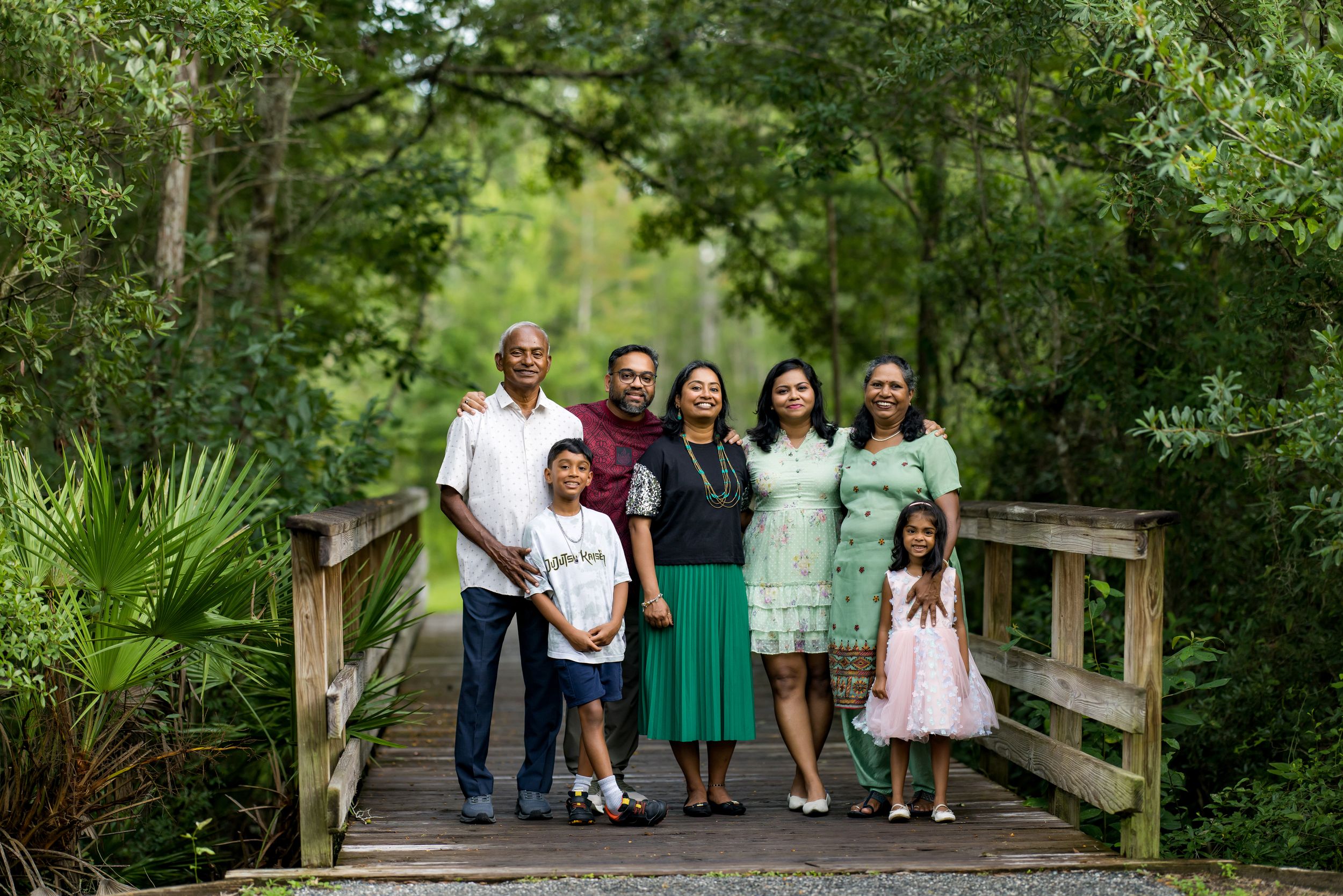 Family of seven posing together on a wooden bridge surrounded by lush green trees in an outdoor park setting.