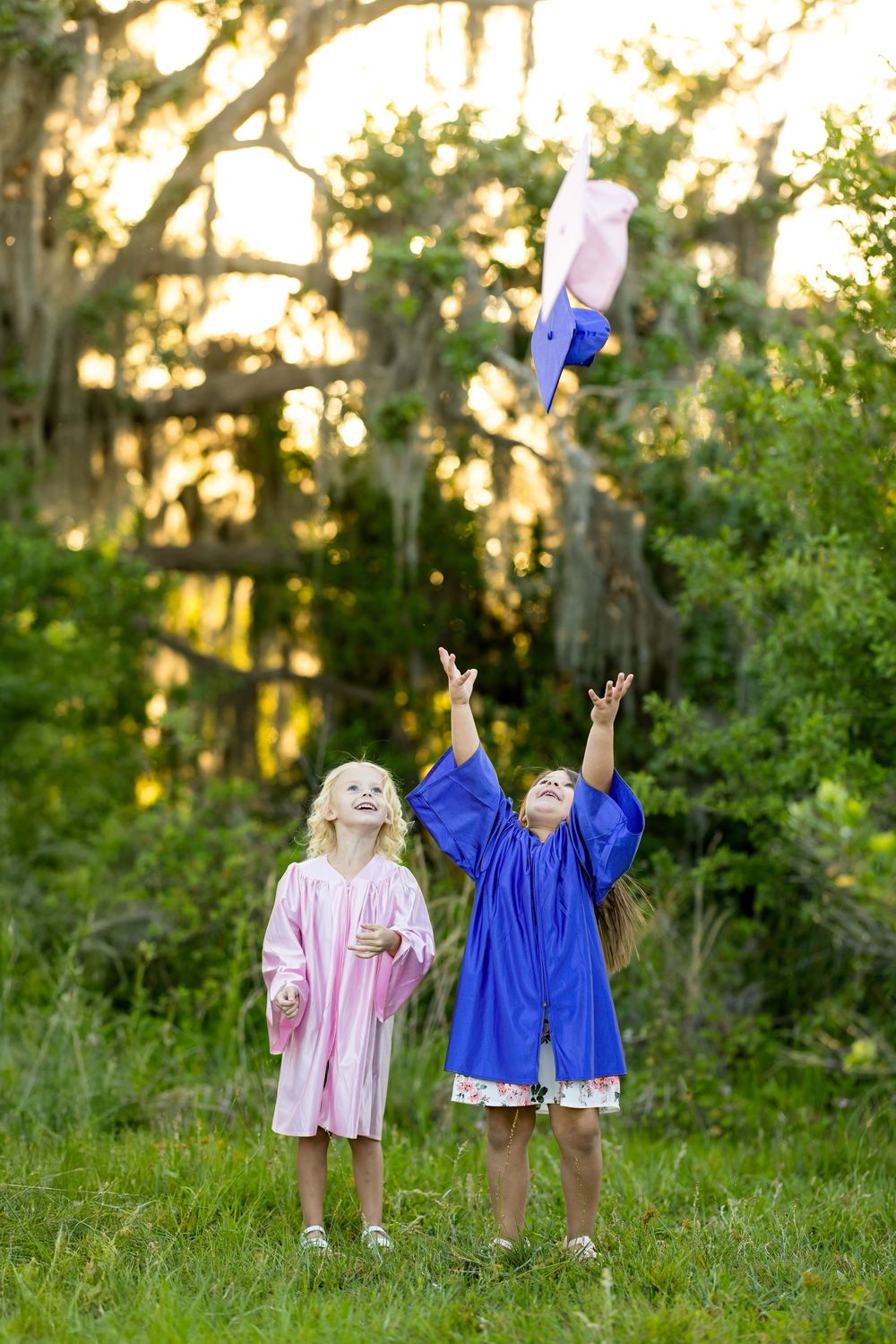 Two children wearing pink and blue graduation gowns play outdoors at sunset in a wooded setting.