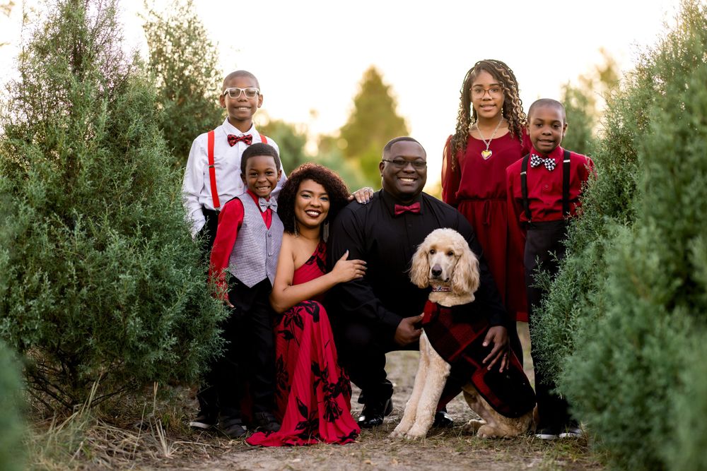 Family poses together in coordinated red and white outfits among Christmas trees at sunset.