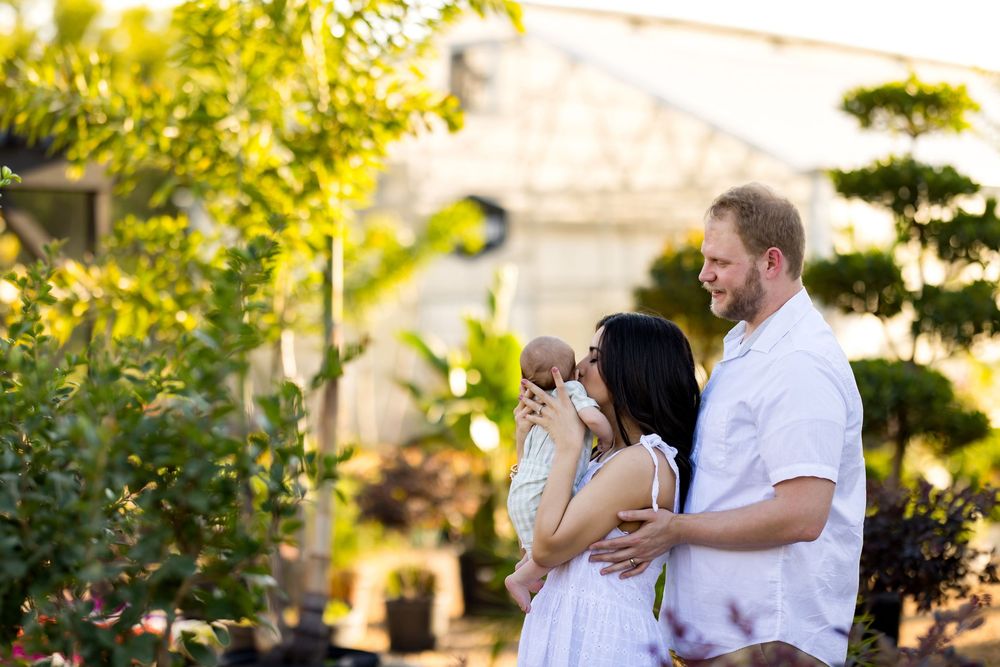 A couple in white outfits holds their newborn baby outdoors near lush greenery and a greenhouse.