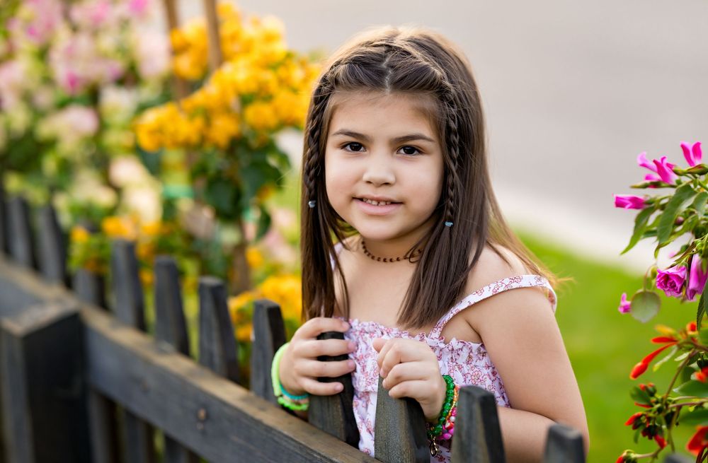 A young girl with braided hair smiles near a wooden fence surrounded by colorful blooming flowers outdoors.