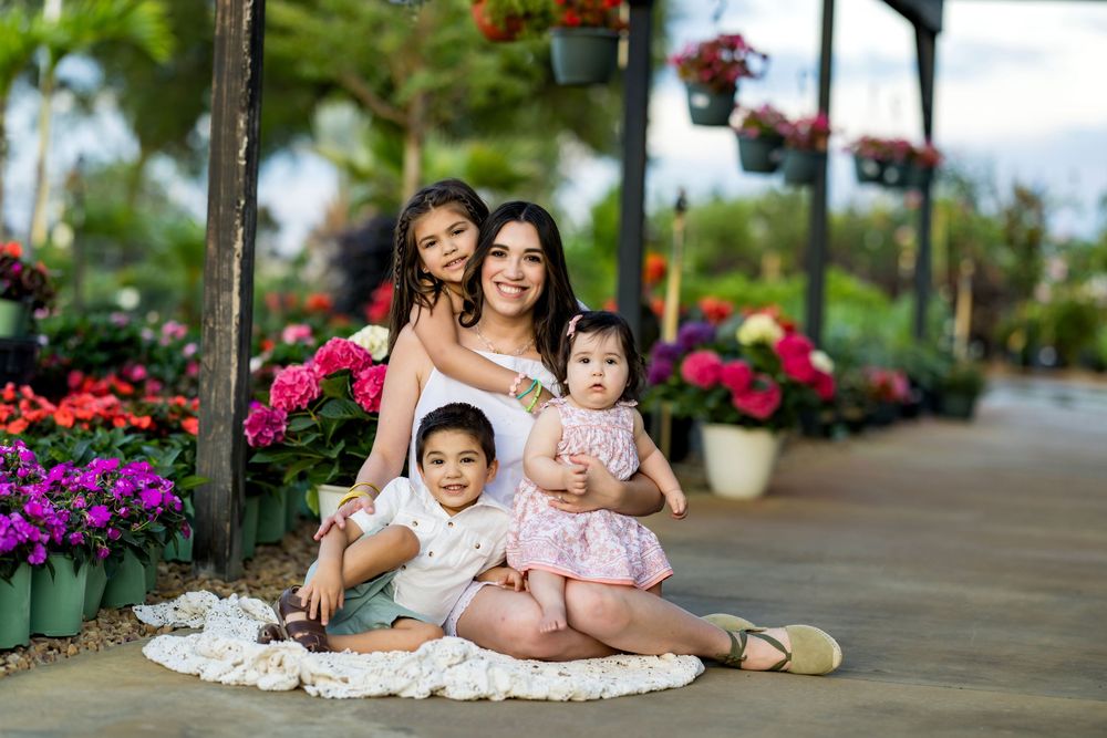 A mother poses with her three young children surrounded by colorful flowers at a garden nursery.