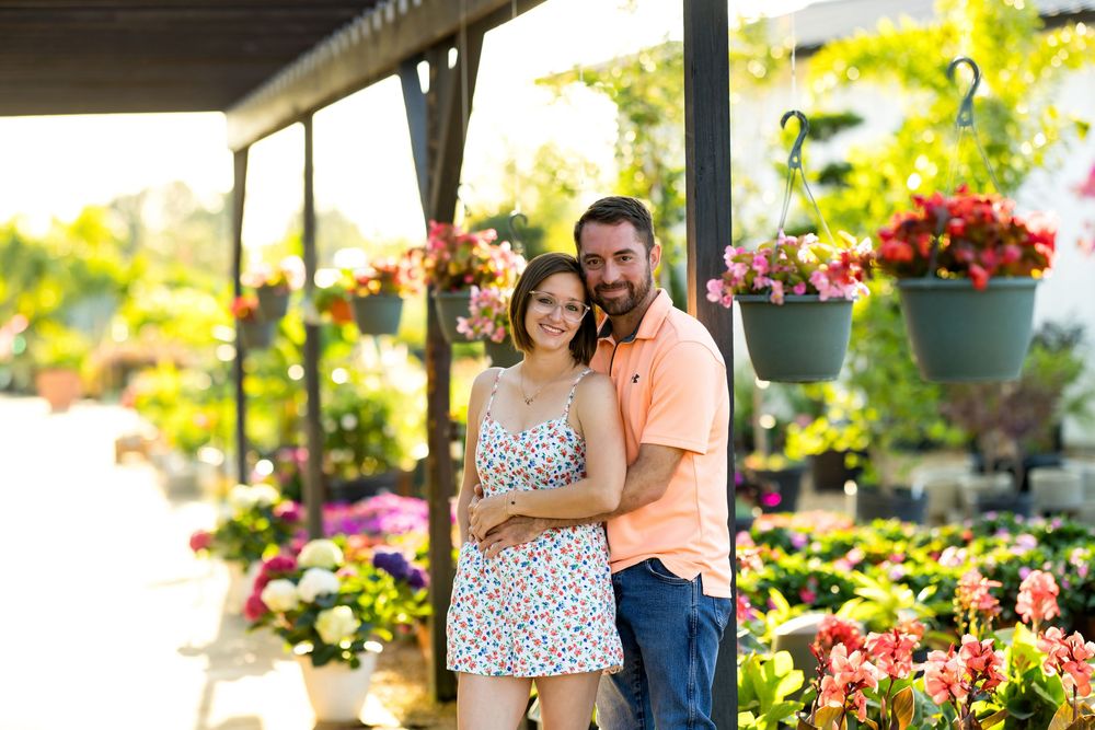 Couple smiling and embracing at a colorful flower nursery with hanging baskets and blooms in background.