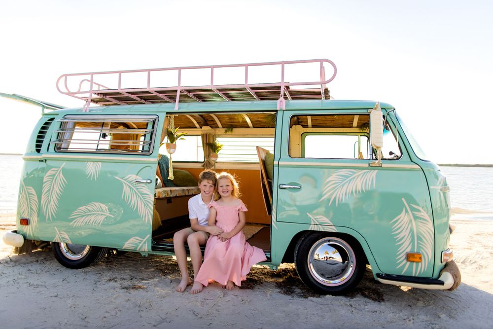 Children sitting in a mint green vintage VW bus with palm leaf designs on a beach.