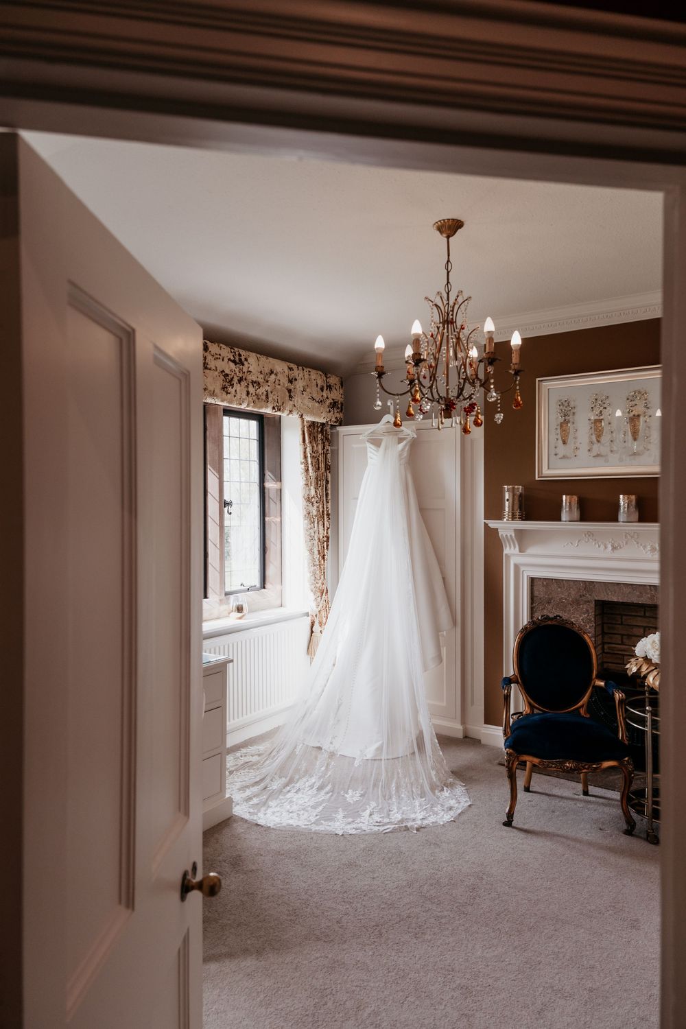 Elegant white wedding dress hanging from a gold chandelier in a classic bridal suite with fireplace.