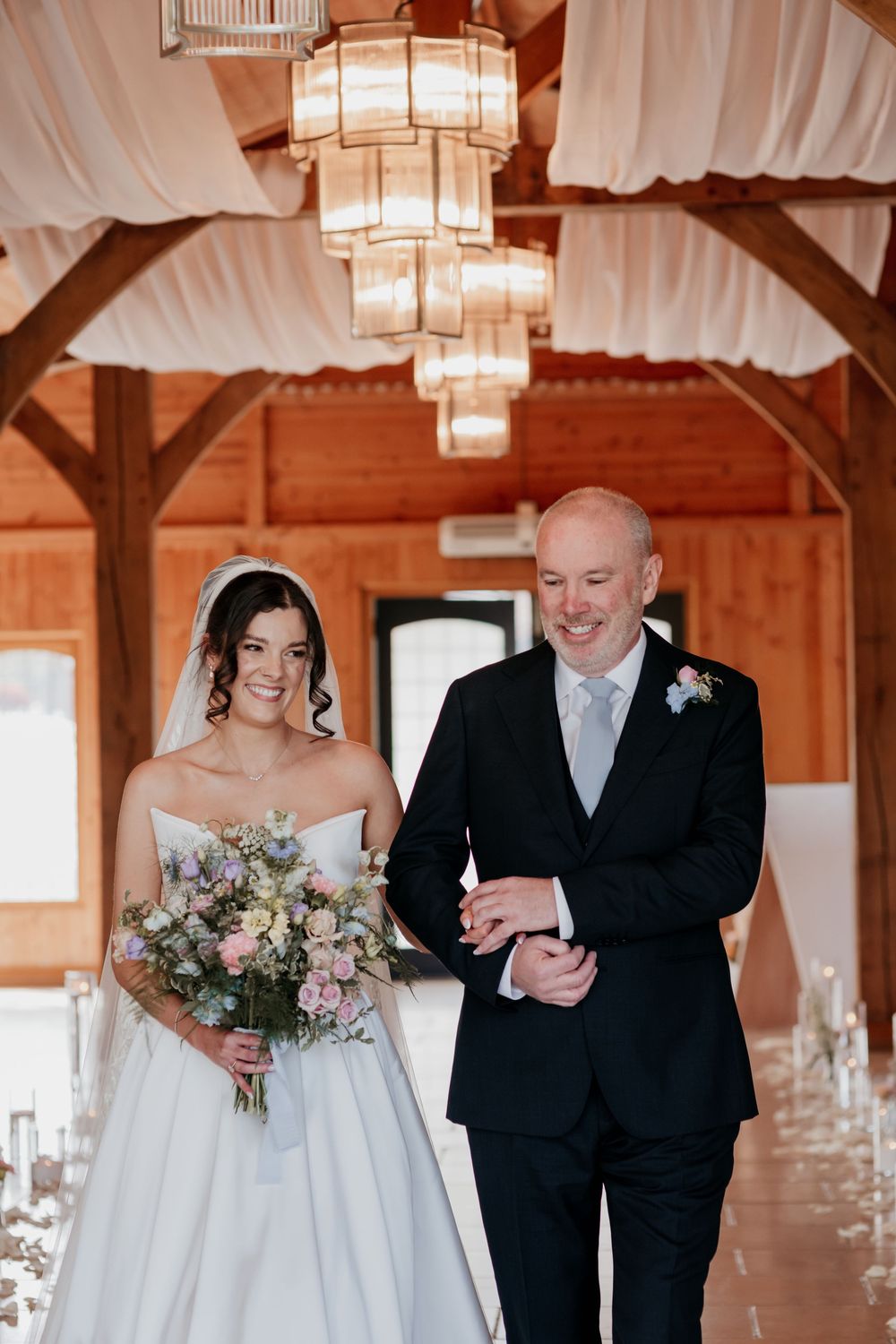 Smiling bride holding floral bouquet walks down aisle with father in rustic wedding venue.