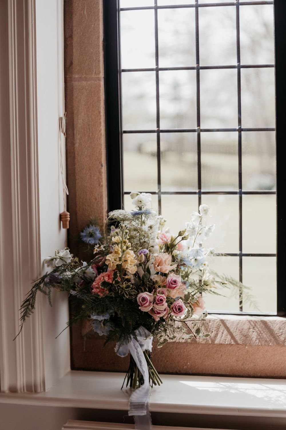 A romantic bridal bouquet with pink roses, blue wildflowers, and white blooms resting on a windowsill with leaded glass windows.