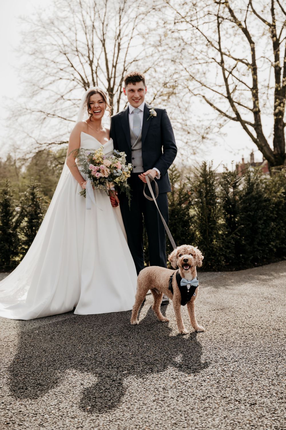 Bride and groom posing with their golden doodle dog wearing a bow tie on their wedding day outdoors.