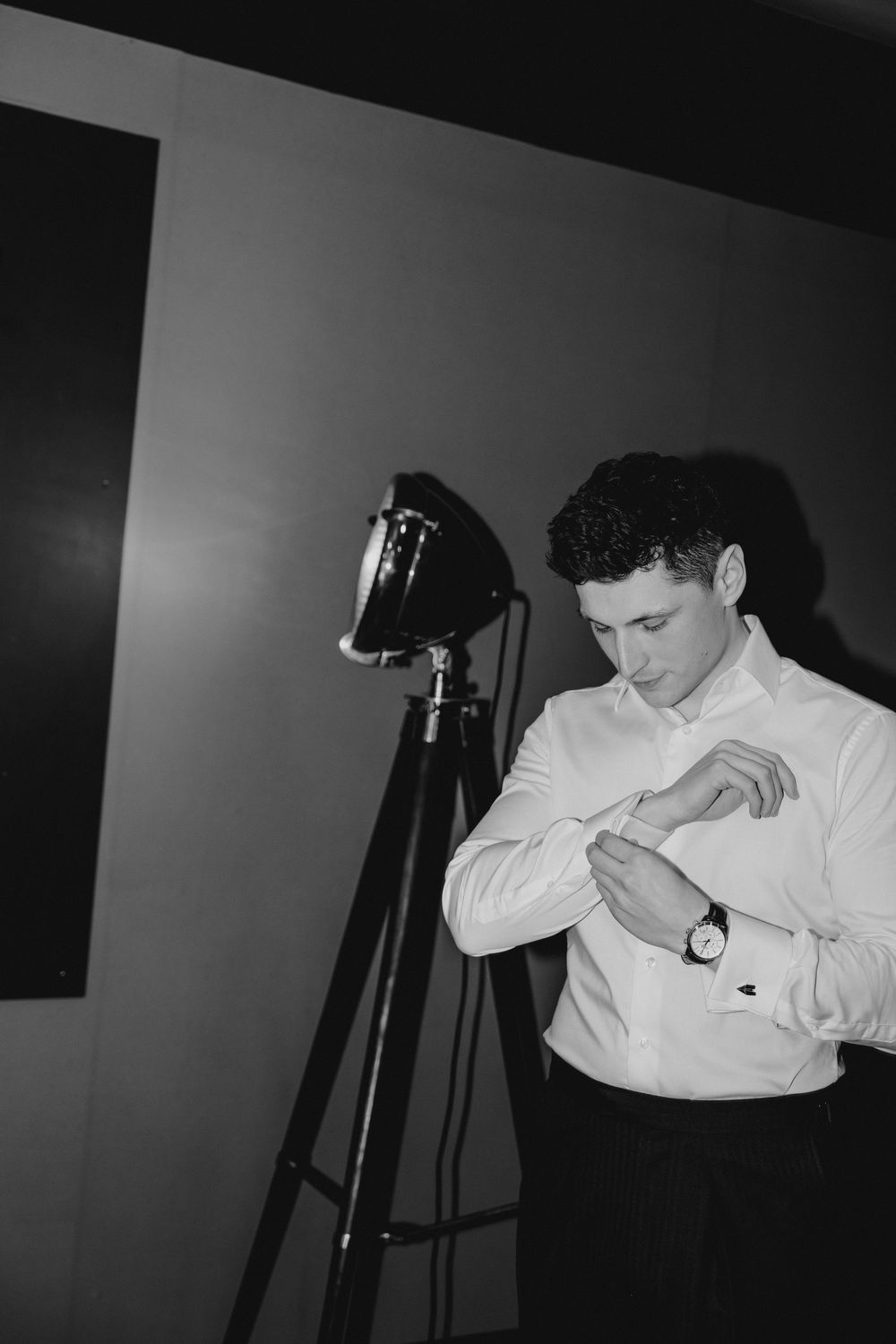 Groom in white dress shirt adjusting cufflinks beside vintage studio light in black and white wedding photo.