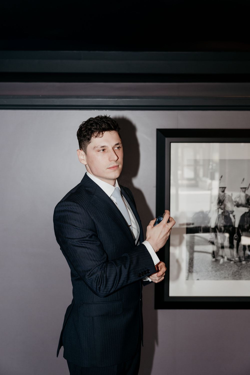Handsome groom in dark suit adjusting cufflinks beside a black and white framed photograph.