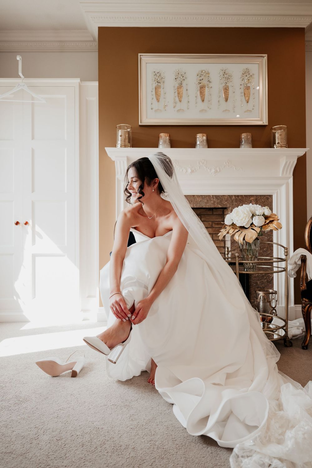 Bride in white gown adjusting her heels before wedding ceremony in elegant room with fireplace.