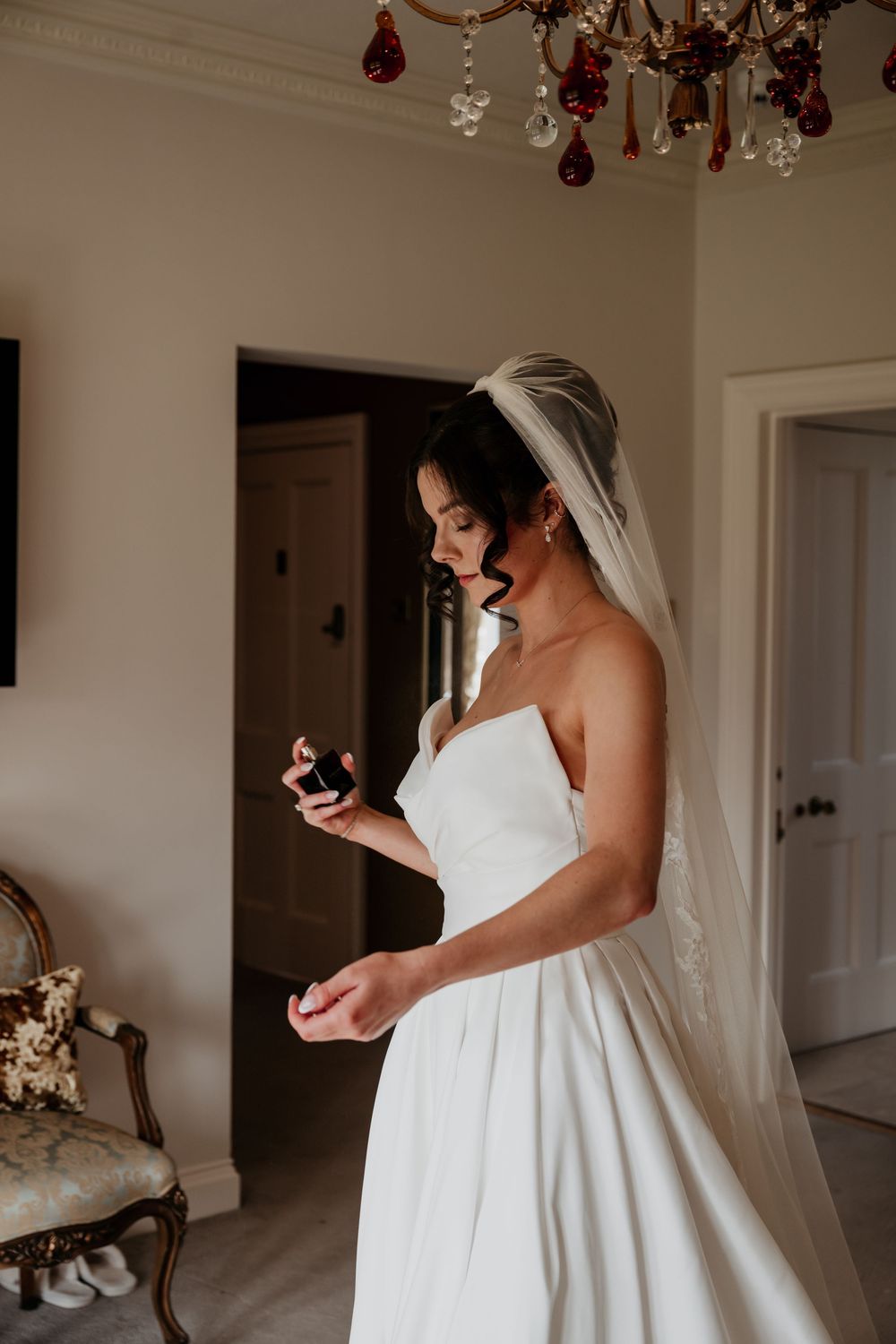 Bride in strapless white gown and veil examines jewelry box in elegant room with chandelier.