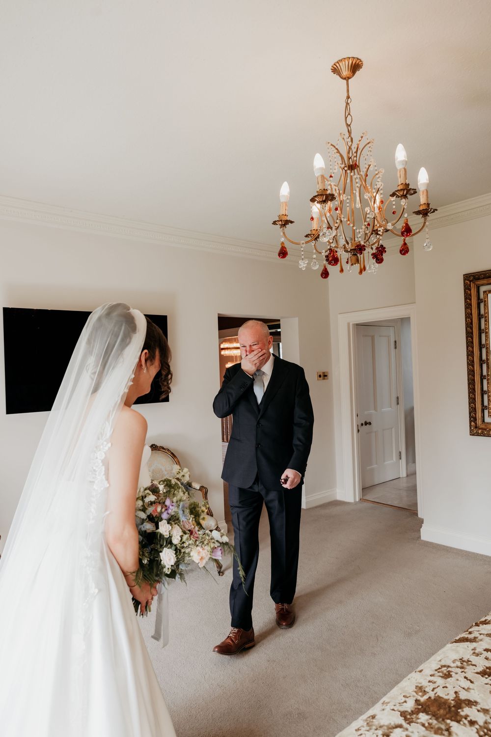 Emotional father wiping tears seeing bride daughter in wedding dress and veil for first time.
