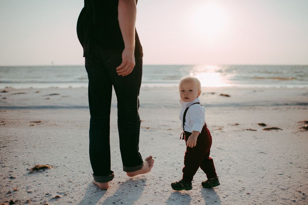 A small child and adult stand barefoot on a sandy beach at sunset with ocean waves in the background.