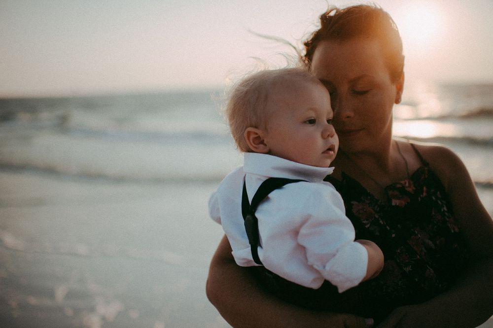 A tender moment captured at sunset as a baby in white and suspenders is held close at the beach. Sarasota best family photographer 2026