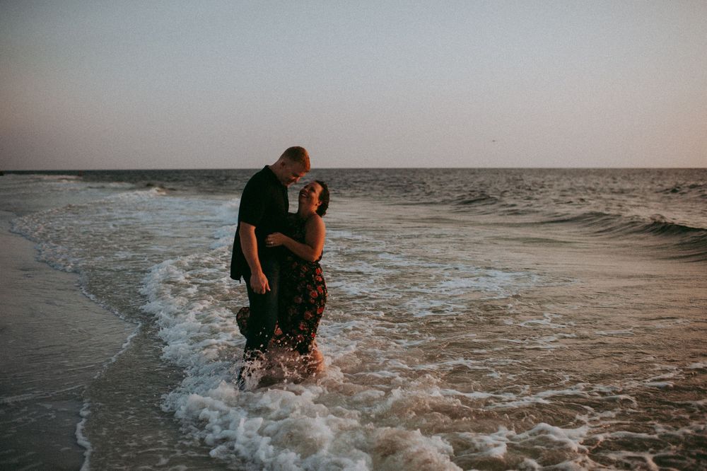 Two people embrace and kiss while standing in shallow ocean waves at sunset.