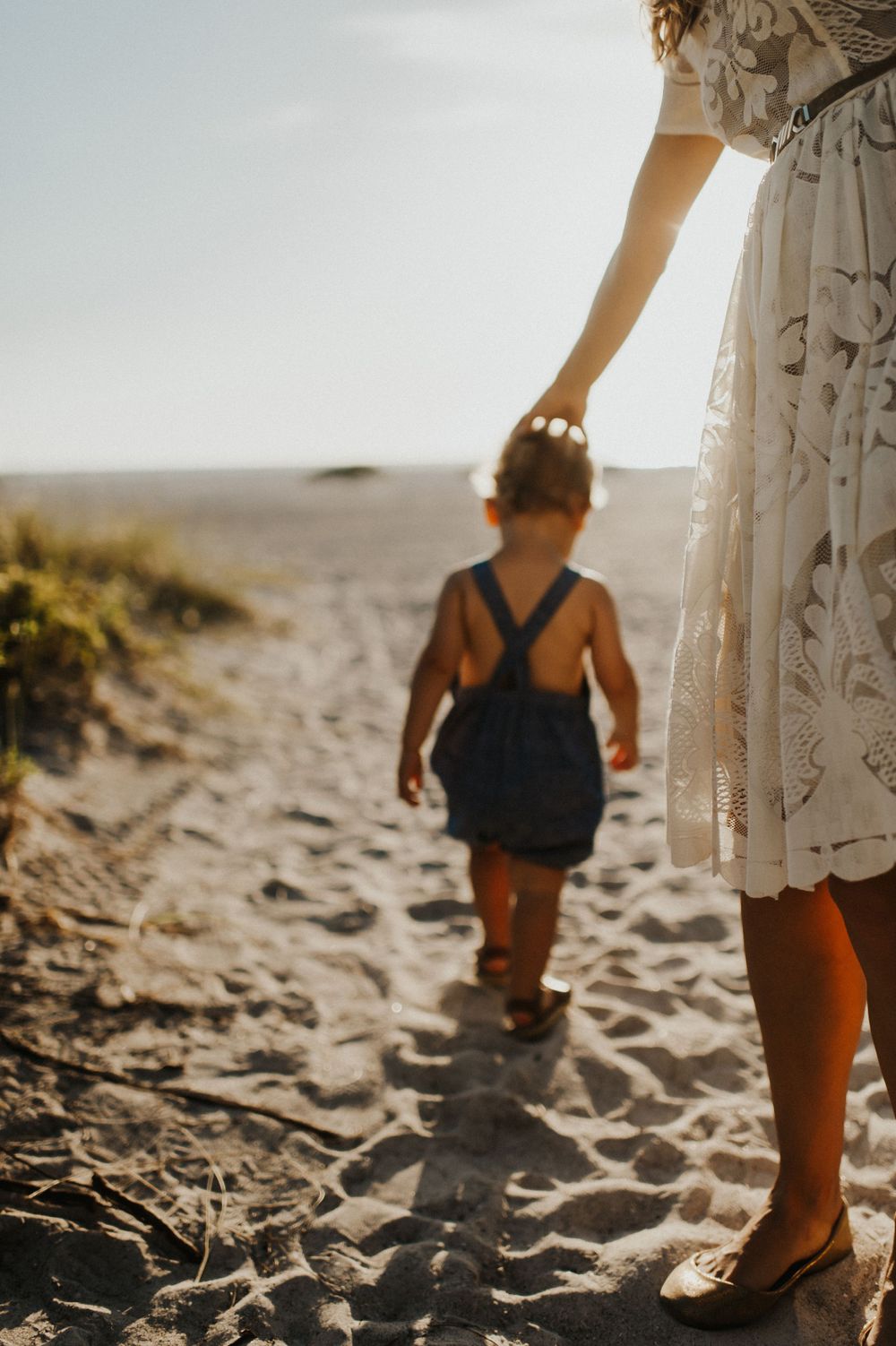A child in overalls walking on a sandy beach path with someone in a white dress. Sarasota Family pictures