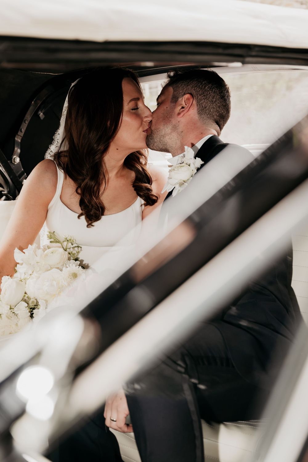 Bride and groom sharing a romantic kiss inside a vintage white car on their wedding day.