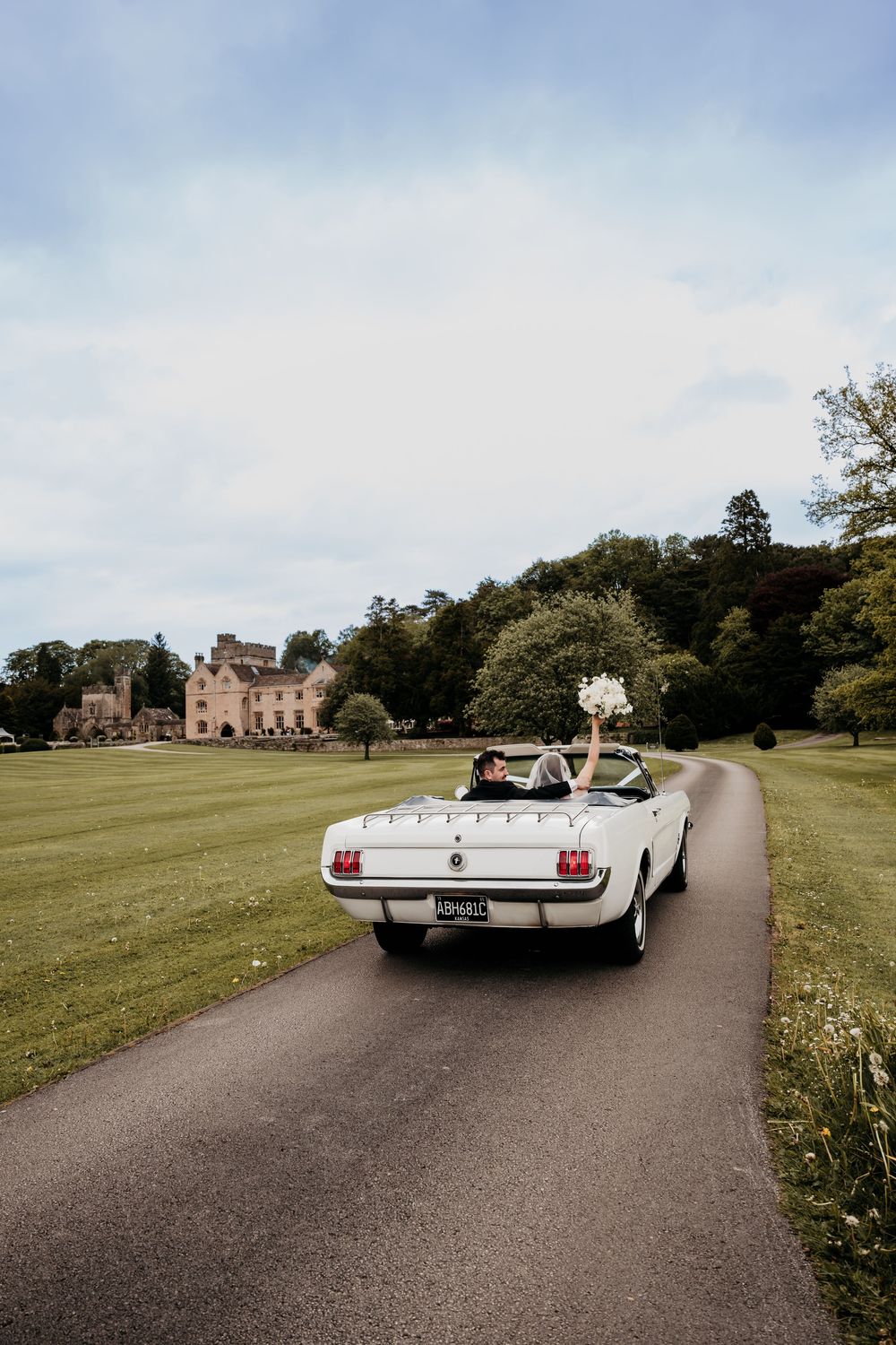 Bride and groom driving away in a white vintage Ford Mustang convertible along a scenic driveway with a grand estate.
