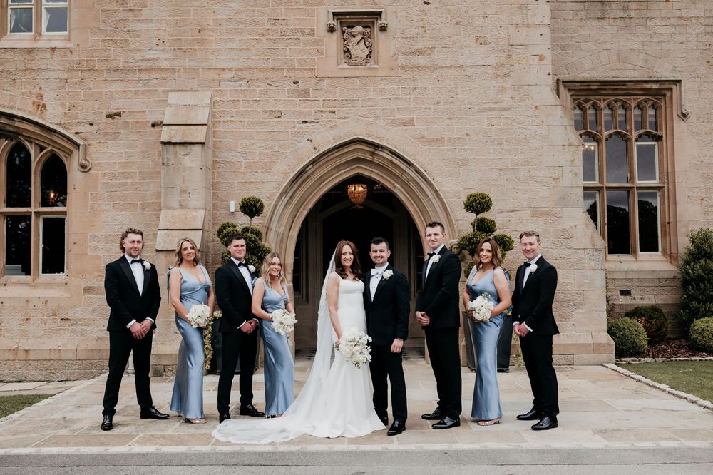 Wedding party posing in front of a grand stone castle entrance in elegant formal attire.