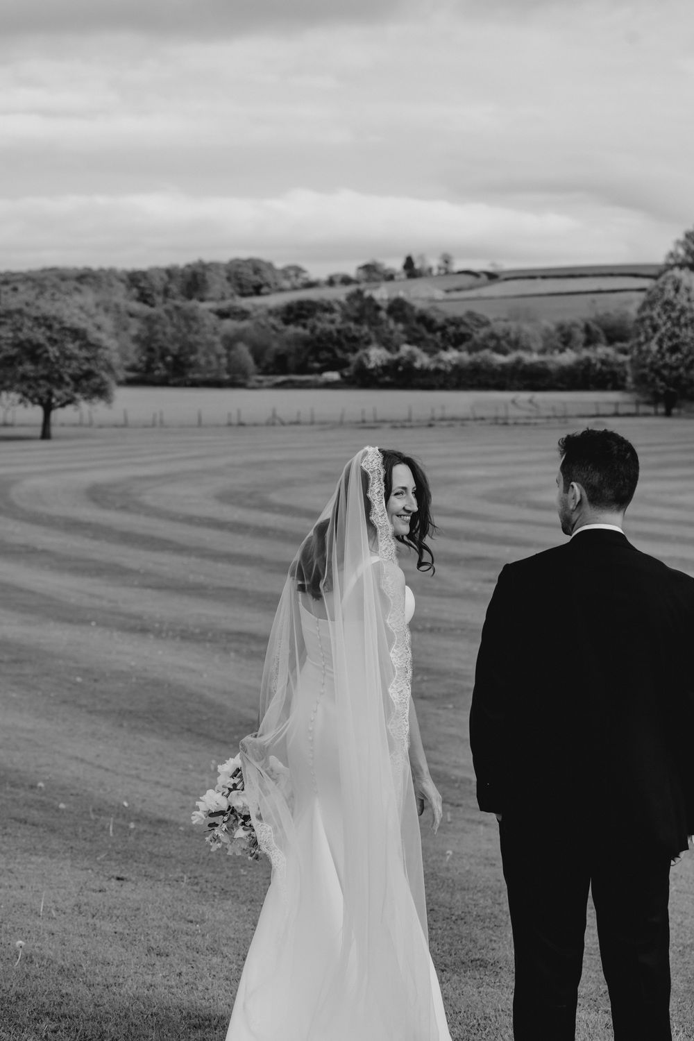 Black and white photo of a smiling bride in a flowing veil and gown holding bouquet beside a suited groom in a scenic countryside.