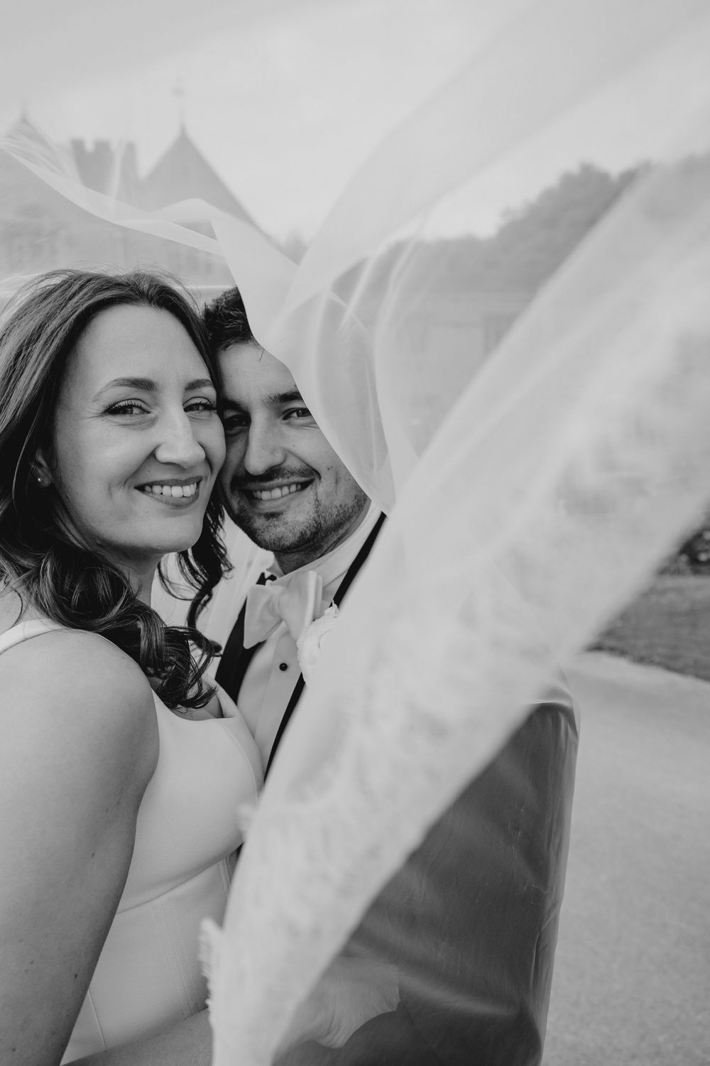 Black and white portrait of smiling bride and groom under flowing veil on wedding day.
