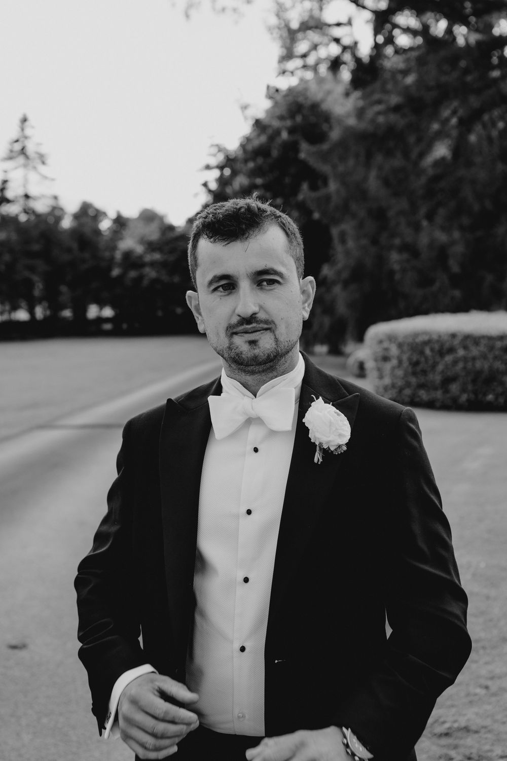 Elegant groom in black tuxedo with white bow tie and boutonniere posing outdoors in a classic black and white wedding portrait.