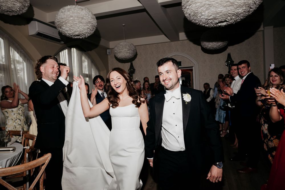 Bride and groom enter wedding reception hall smiling, surrounded by applauding guests and decorative hanging sphere centerpieces.