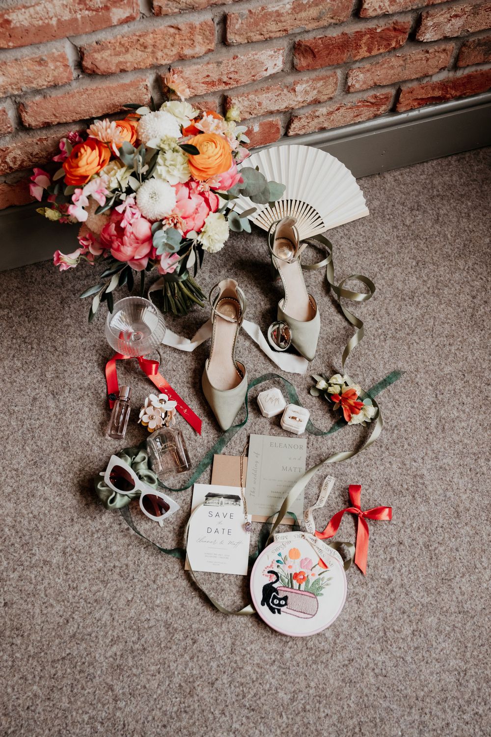 Elegant wedding flat lay with bridal heels, colorful bouquet, save the date cards, rings, and accessories on carpet.