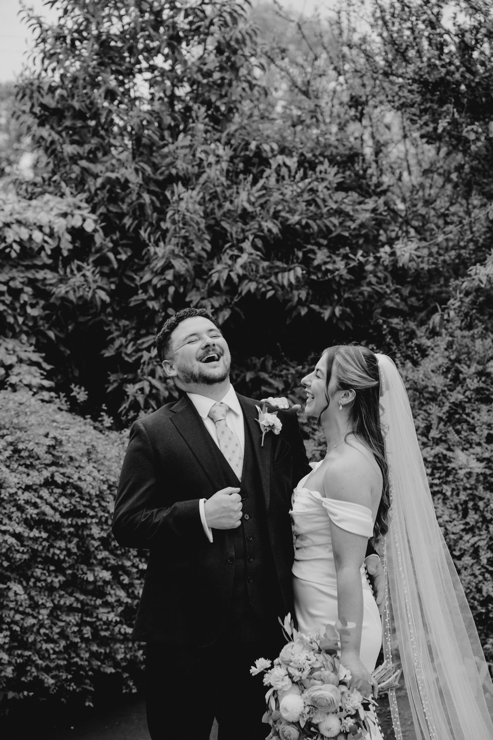 Black and white photo of a laughing bride and groom in formal wedding attire, standing before lush garden greenery.