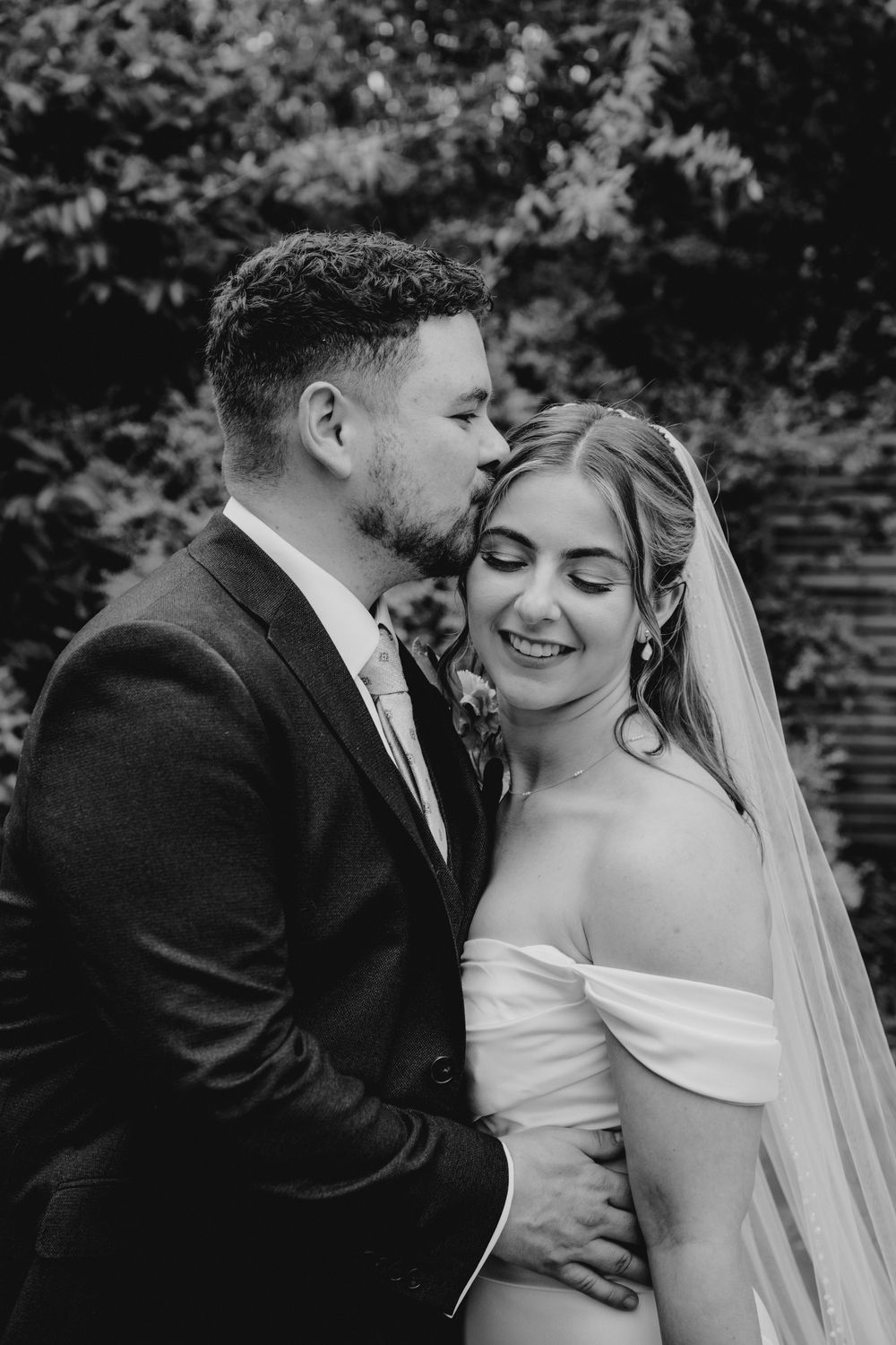 Black and white wedding photo of groom kissing bride on cheek, bride smiling in off-shoulder gown and veil.