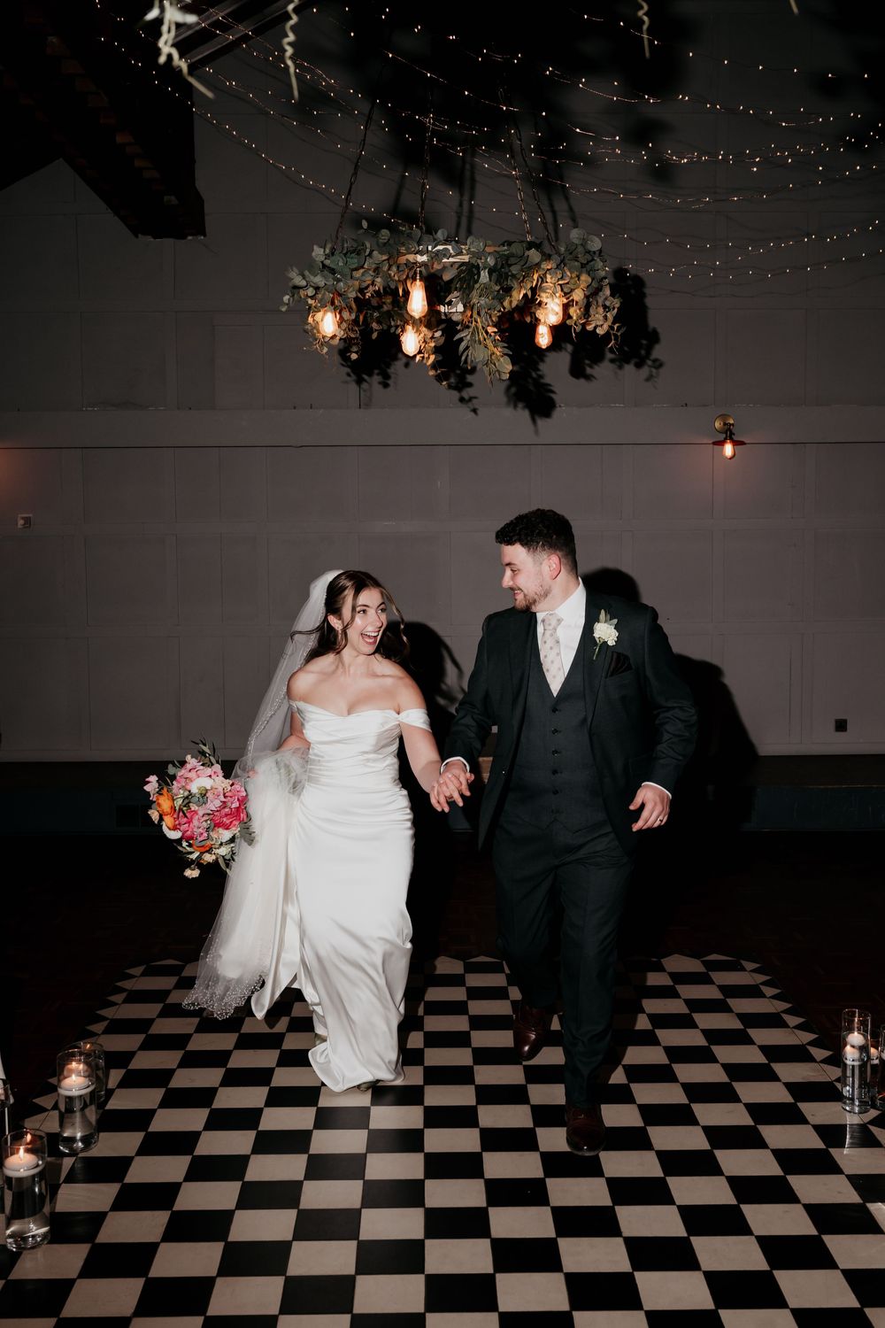 Bride and groom walk hand-in-hand on checkered dance floor under floral chandelier with fairy lights at elegant wedding reception.