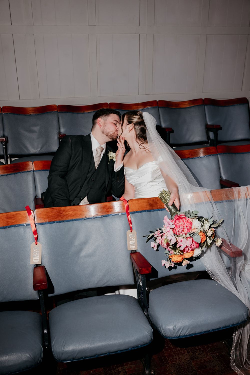 A bride and groom share a kiss while seated in vintage theater chairs, the bride holding a colorful pink and orange bouquet.