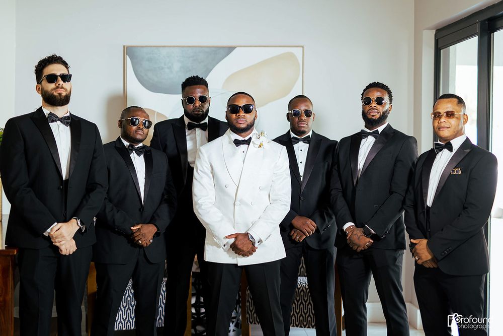 Groomsmen in formal black tuxedos pose with groom in white tuxedo for wedding photo.