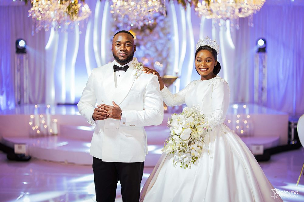 Wedding couple in white attire stand on reflective floor with illuminated backdrop.