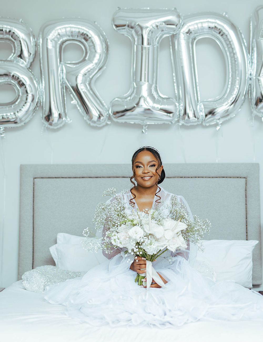 A bridal portrait with silver BRIDE balloons and white floral bouquet on a bed.