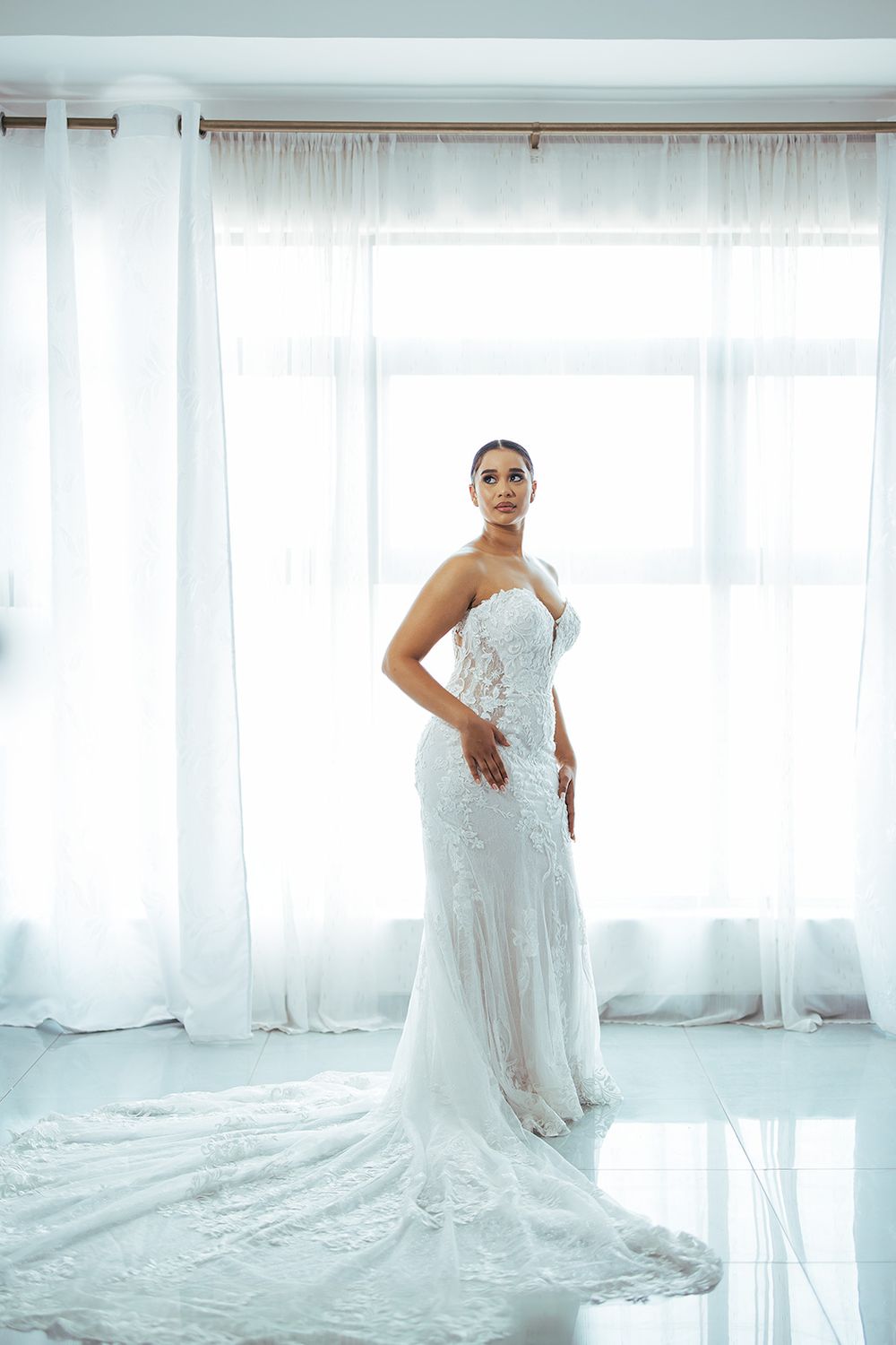 An elegant bride in a fitted lace wedding gown poses against sheer white curtains.