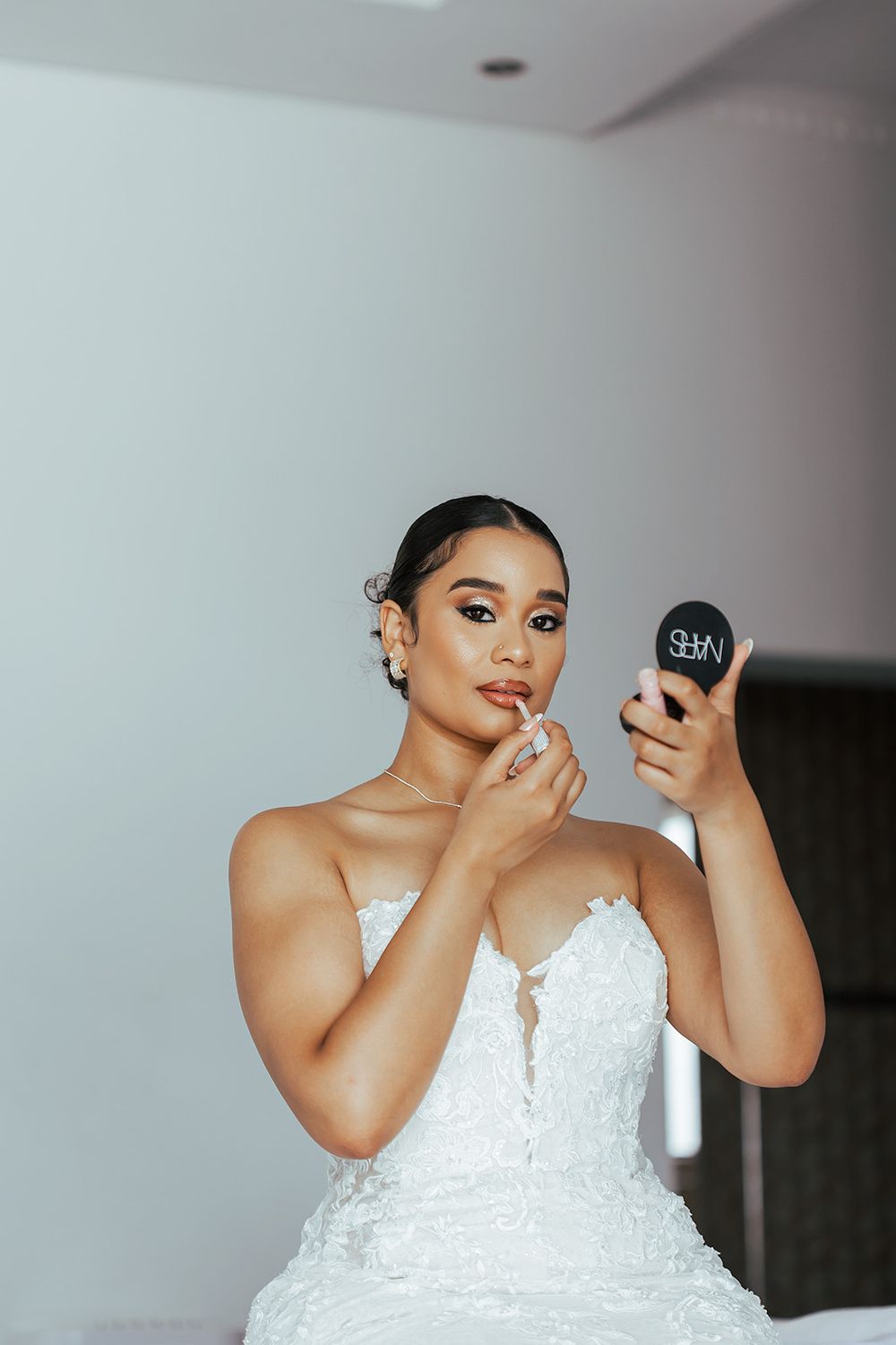 Bride in white lace wedding dress applies makeup while looking in compact mirror.