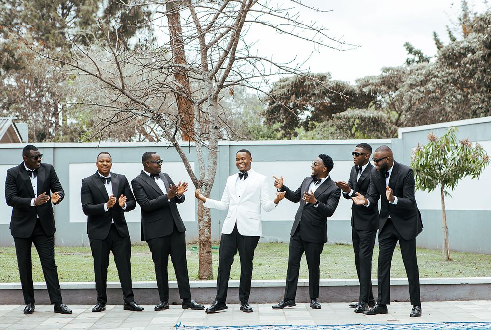 Groomsmen in black suits pose casually outdoors with groom in white jacket.