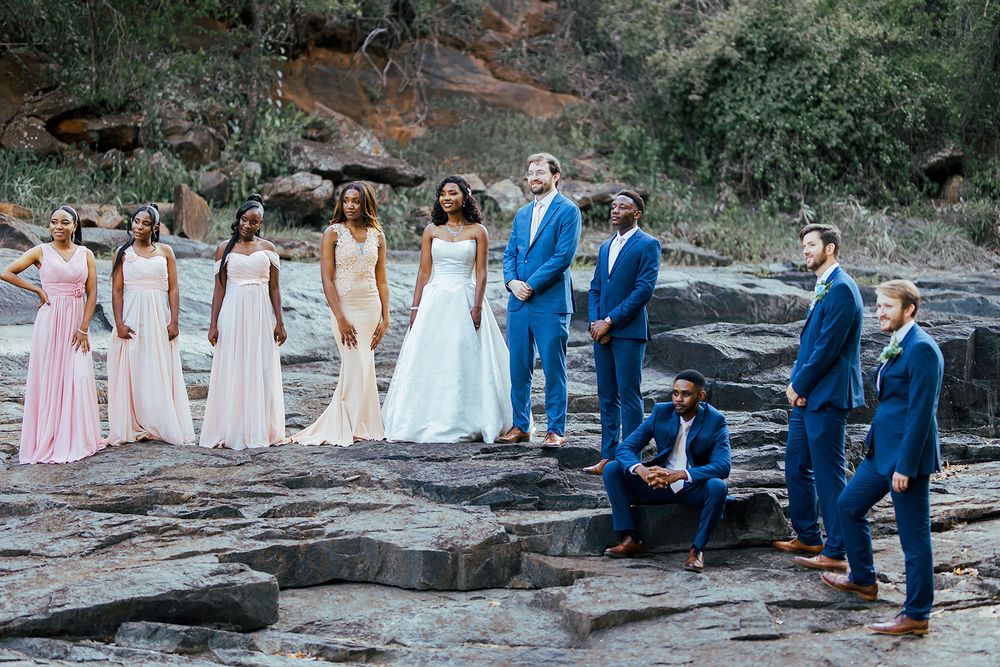 Wedding party in blue suits and pink dresses poses on rocky outdoor terrain.