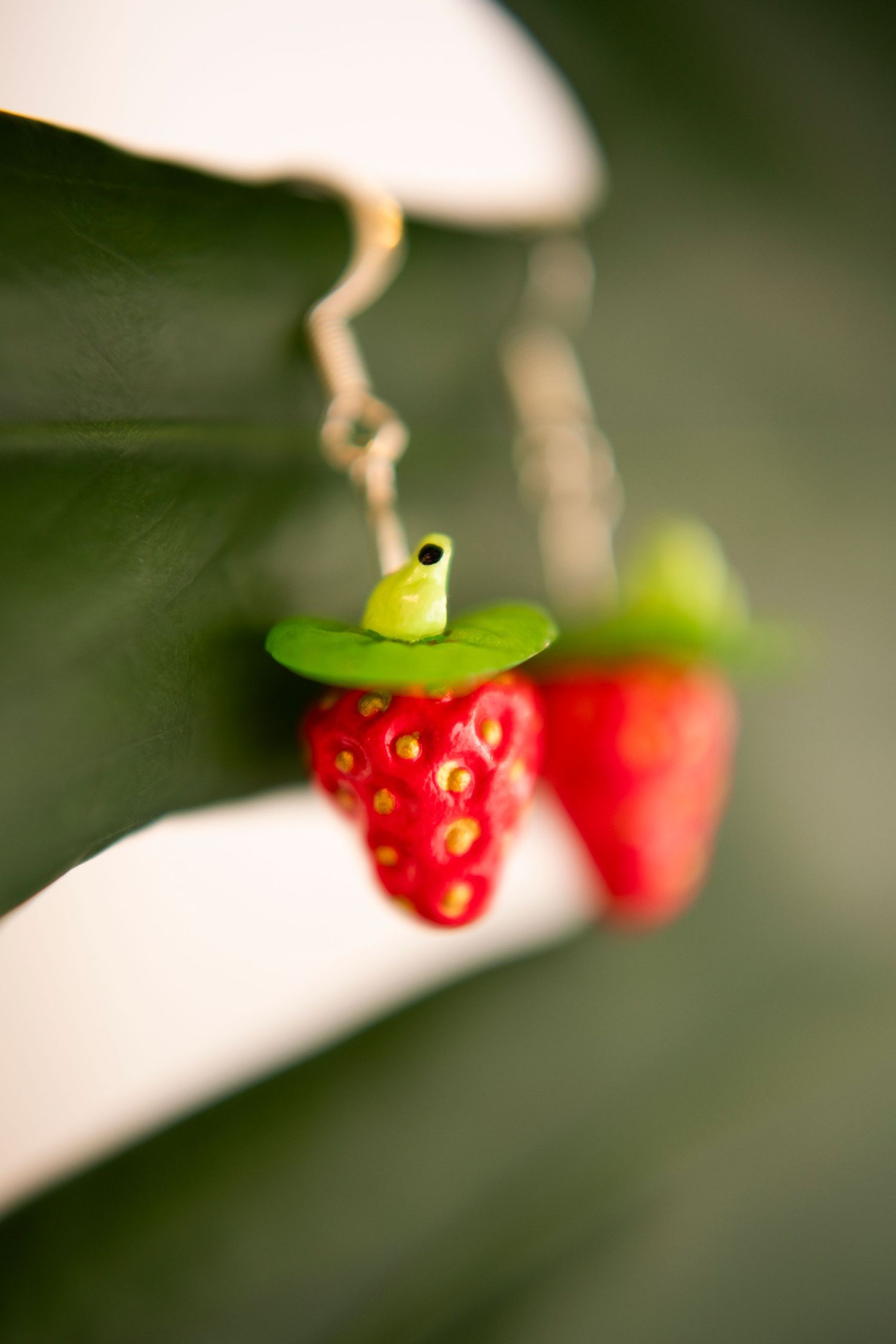A delicate strawberry-shaped earring with a green leaf accent hangs from a gold chain against a blurred background.