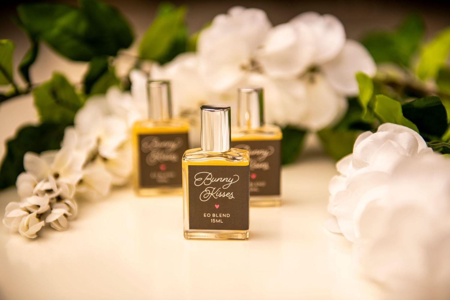 Three gold perfume bottles with white floral elements displayed against a soft white background with green foliage accents.