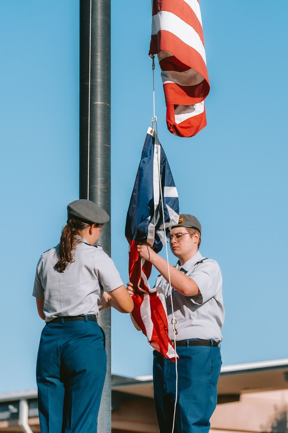Two people in uniform raising American flag on flagpole against blue sky.