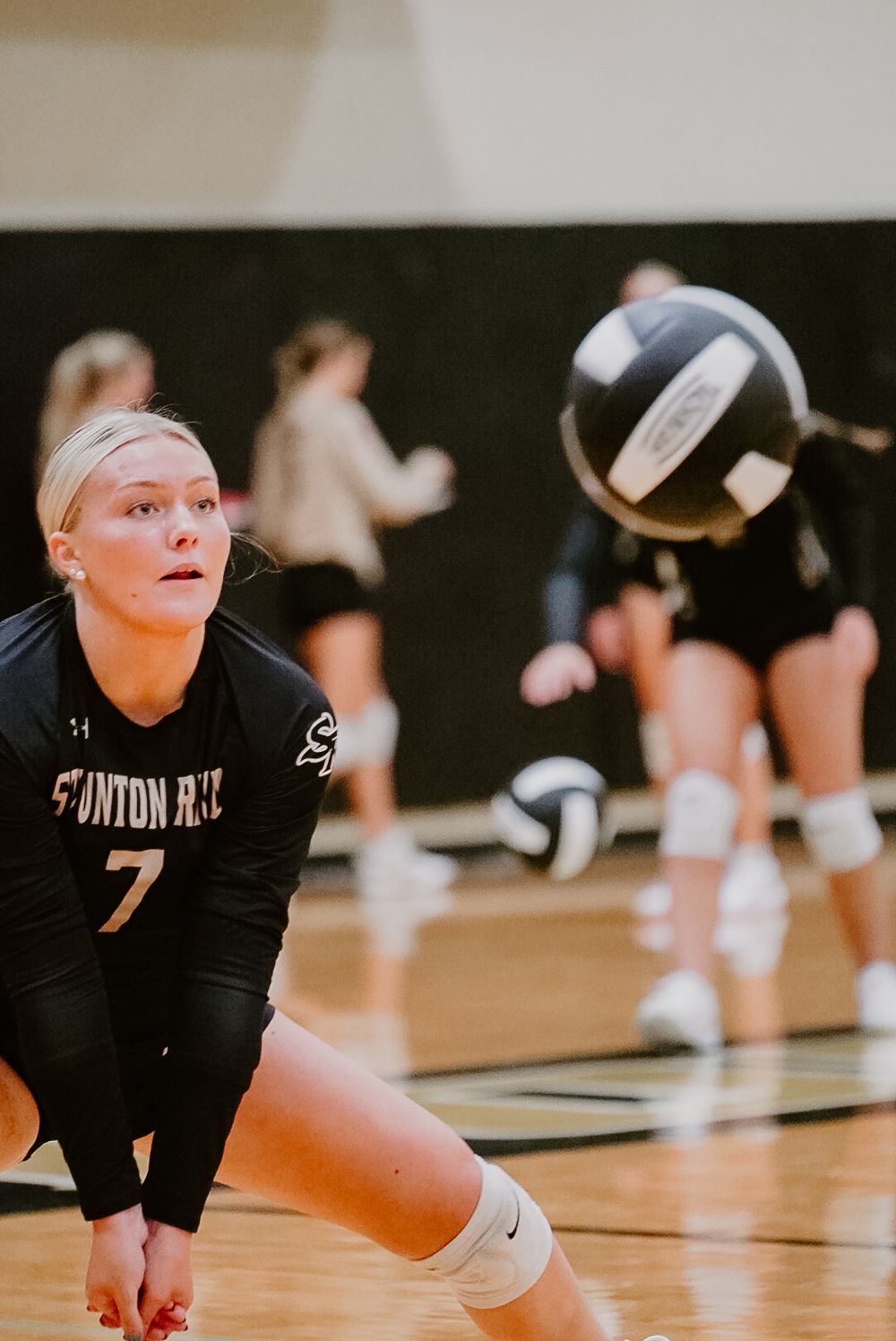 Volleyball player in black Clinton jersey number 7 prepares to receive serve during indoor match.