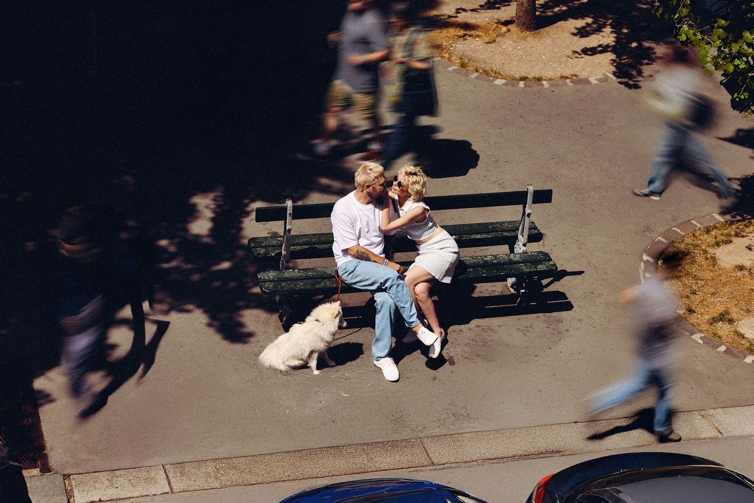 People sit and relax on benches in a sunny park area while others walk past in the background.